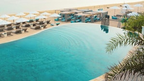 A poolside area with lounge chairs and umbrellas, a sandy beach, and a clear blue pool. - Infinity8 Bali (Jimbaran)