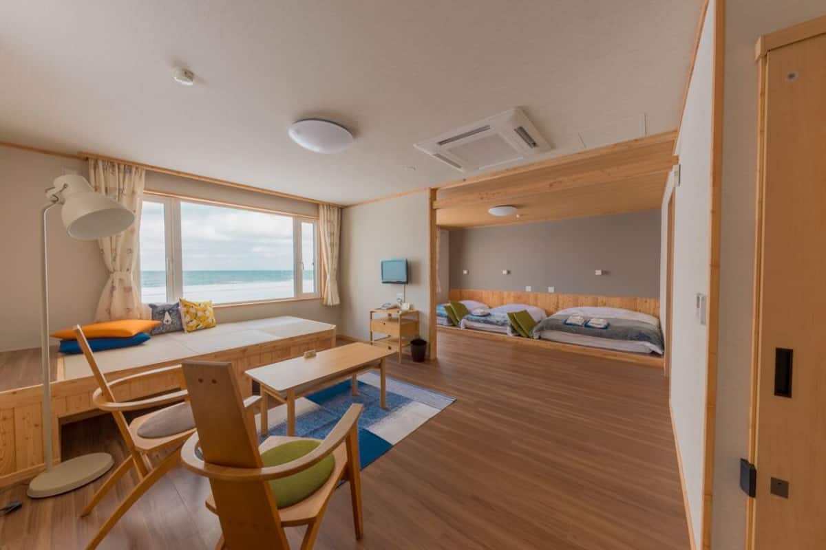 A room with a wooden floor, a desk with a chair, a bed with pillows, a window with curtains, and an air conditioning unit.