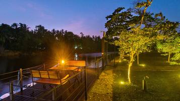 A wooden deck with a bench overlooking a body of water, surrounded by trees and illuminated by ambient lighting.