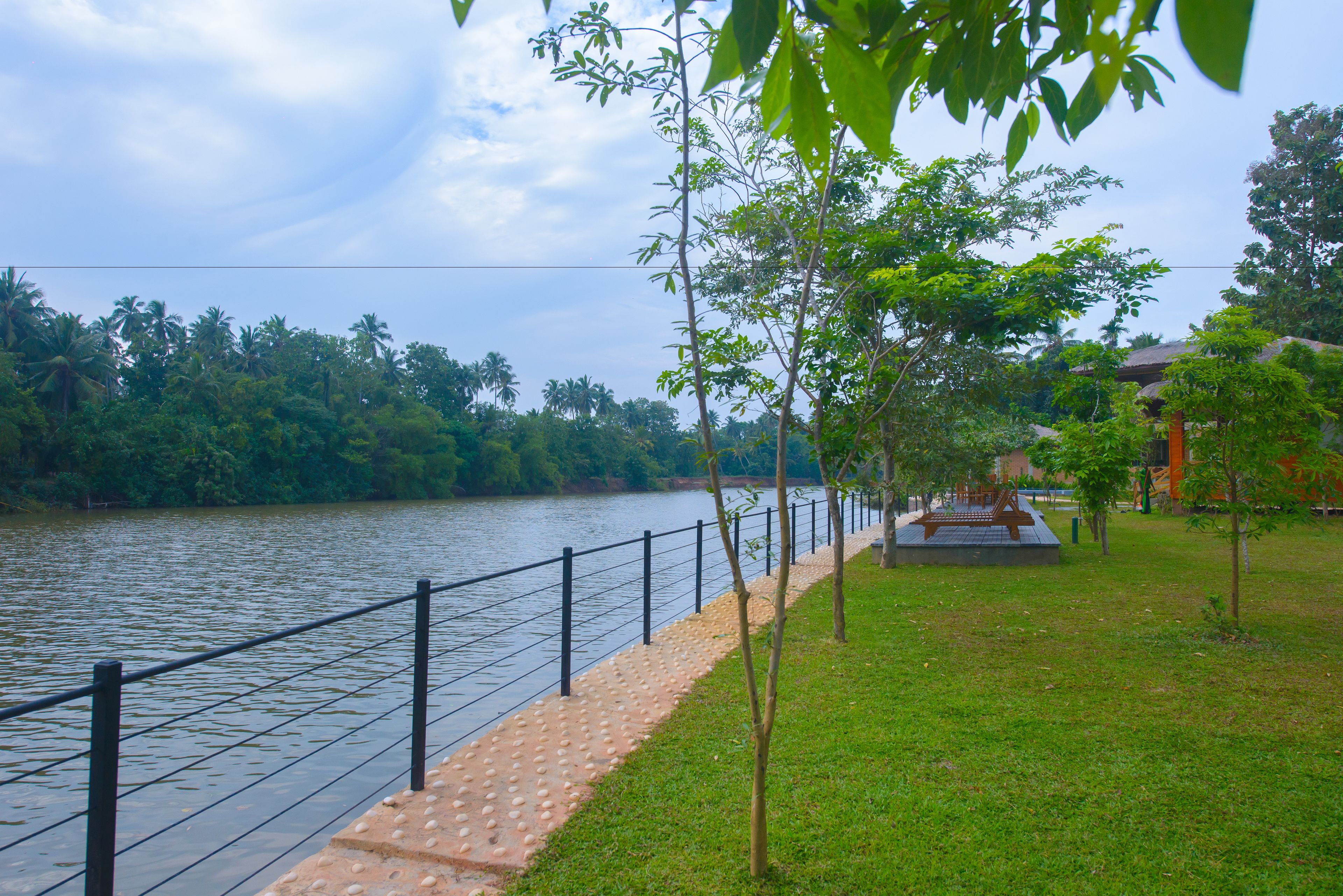 Un paseo junto al río con valla, árboles y una pequeña zona de parque.