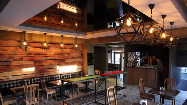 Un restaurant moderne avec un mur en bois, de nombreuses tables et chaises, et des luminaires géométriques suspendus.