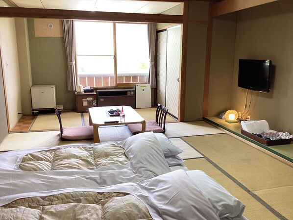 A traditional Japanese-style room with tatami flooring, a futon bed, a small table with a chair, a television, and a kitchenette area.