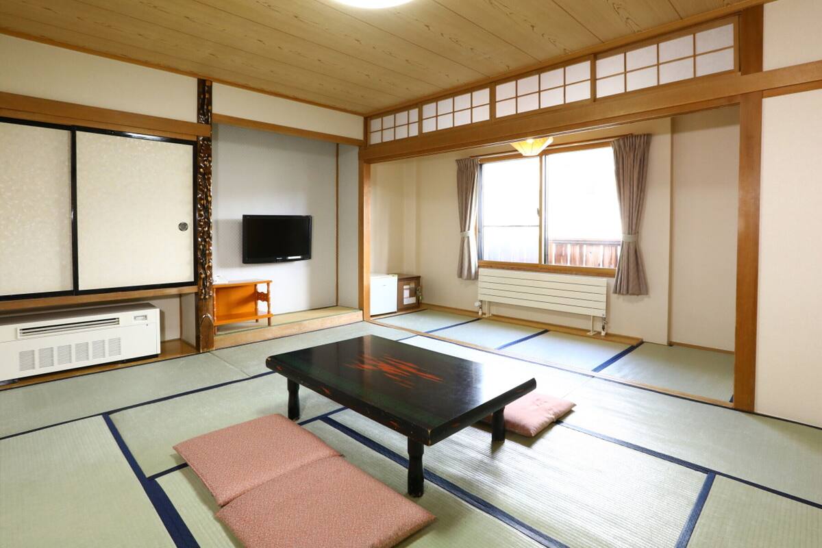 Une pièce de style japonais traditionnel, avec un sol recouvert de tatamis, une table basse, des coussins, un téléviseur et une fenêtre avec des rideaux.