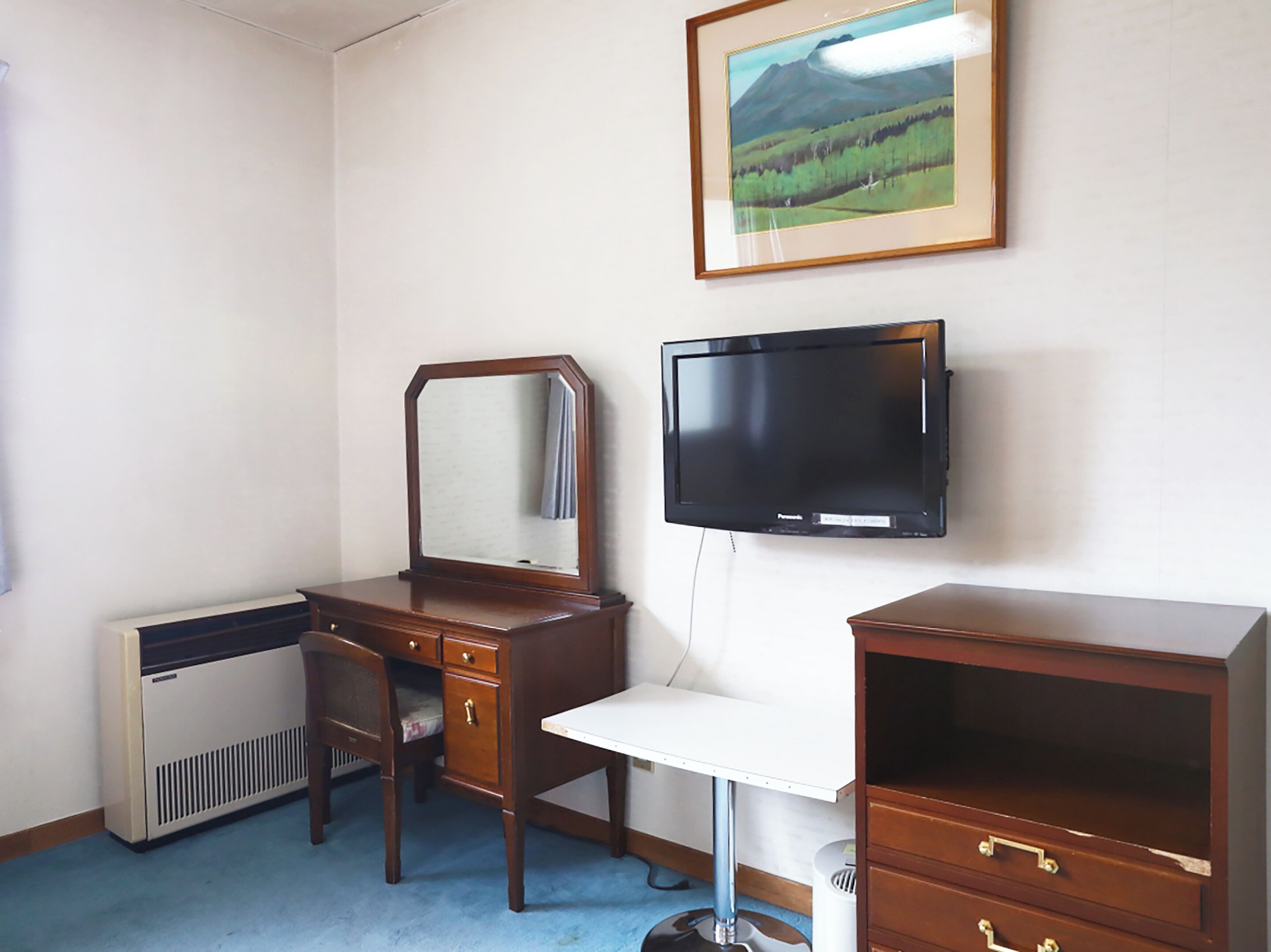A room with a wooden desk, a mirror, a television, and a framed landscape painting on the wall.