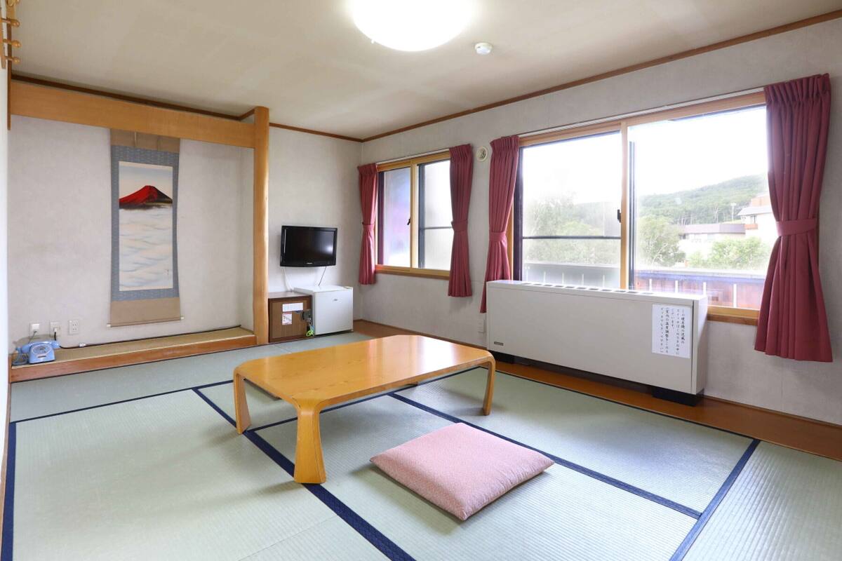 Une pièce de style japonais traditionnel, avec un sol recouvert de tatamis, une petite table en bois, un téléviseur et une fenêtre ornée de rideaux rouges.