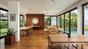 A spacious dining area with wooden tables and chairs, large windows offering views of a swimming pool and greenery, and a ceiling fan. - Ubuntu Beach Villas By Reveal (Kamburugamuwa)