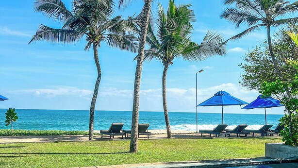 A clear blue pool with lounge chairs and umbrellas, palm trees, and a view of the ocean. - Ubuntu Beach Villas By Reveal (Kamburugamuwa)