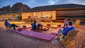 A desert scene with a group of people seated on striped mats and wicker chairs, a stage with musical instruments, and a building with a striped awning. - Mohammed Mutlak Camp (Wadi Rum)