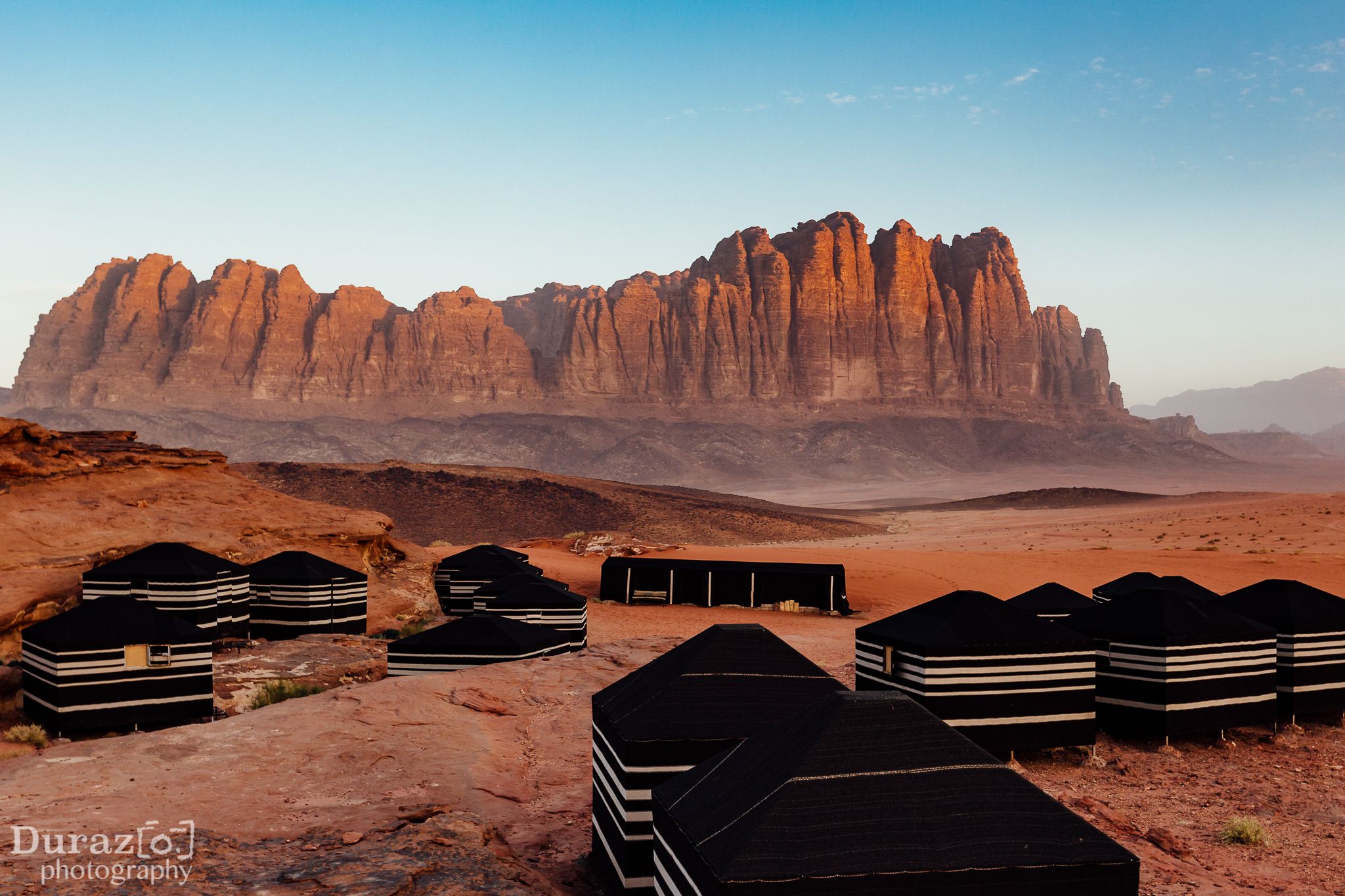 A desert landscape with a large rock formation in the background and several black tents in the foreground.