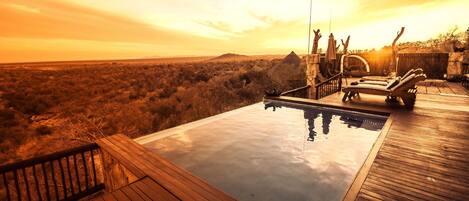 A wooden deck with a swimming pool, surrounded by a natural landscape and a sunset.