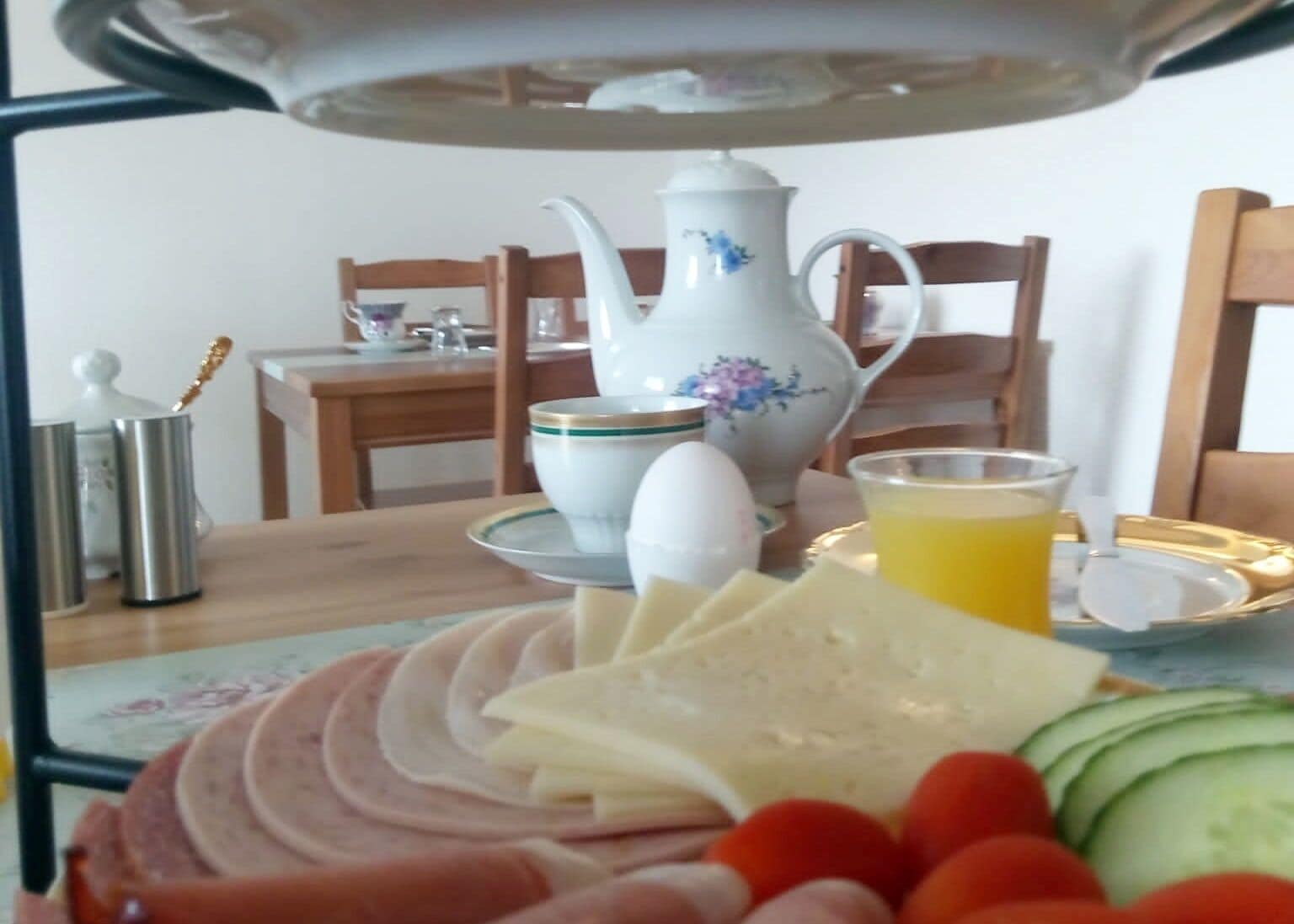 Disposição para o café da manhã com presunto fatiado, queijo, tomates e pepinos em um prato, um bule, uma xícara e um copo de suco de laranja sobre uma mesa.