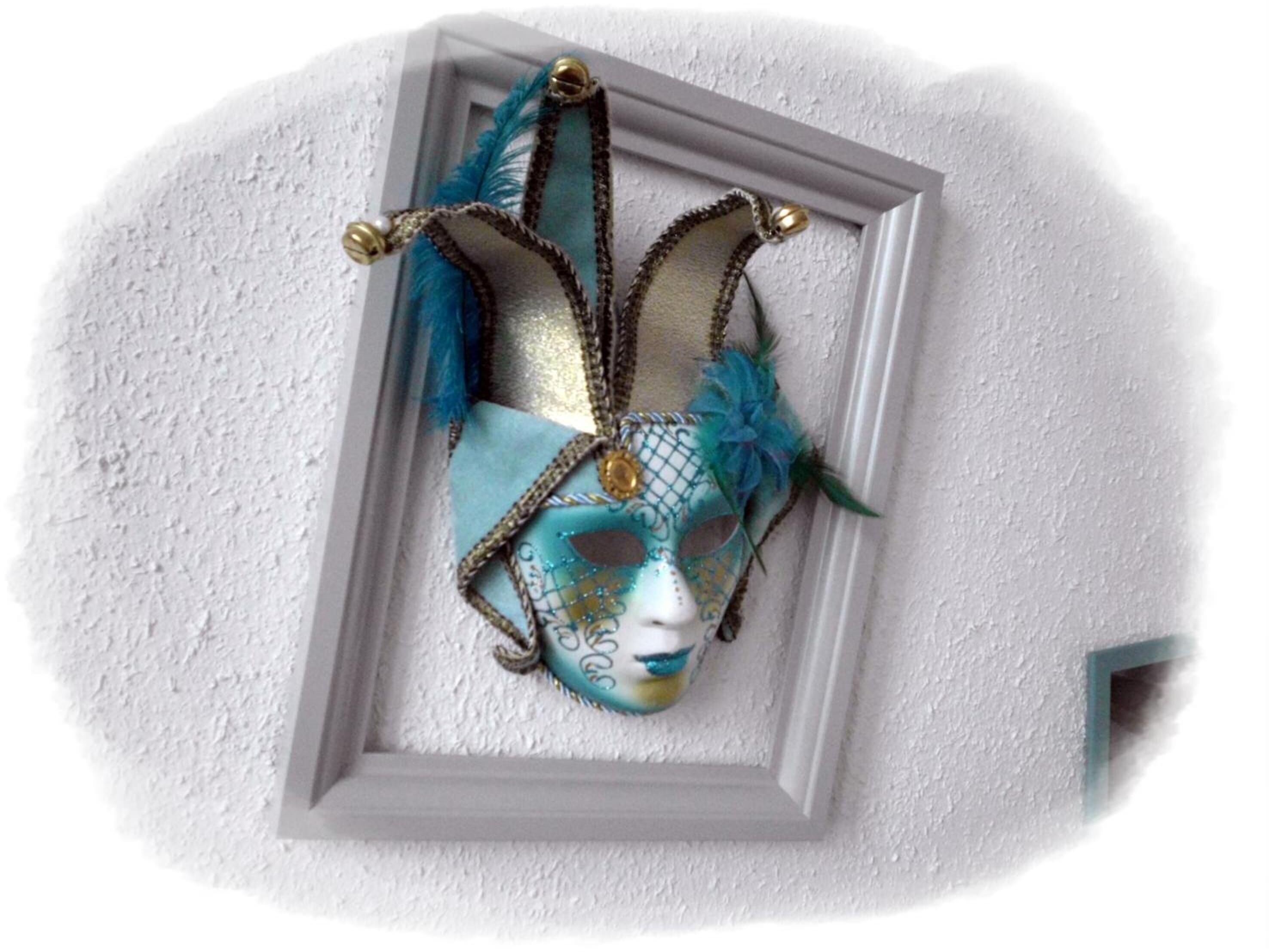 Un masque décoratif orné de plumes et de bijoux, exposé dans un cadre blanc sur un mur à texture.