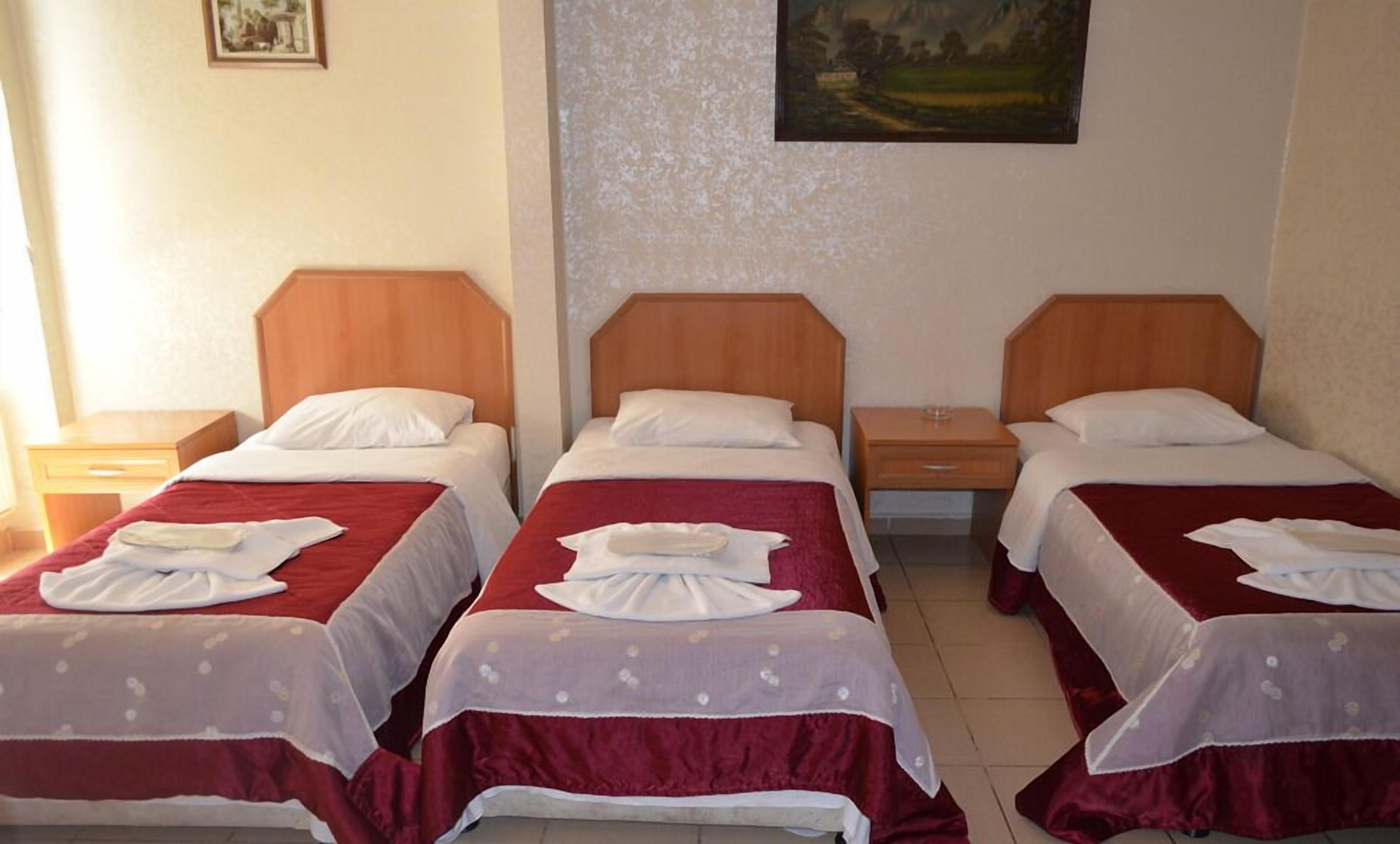 Habitación de hotel con tres camas individuales, cada una con cabecero de madera, almohadas blancas y colchas rojas. Hay mesitas de noche de madera con un vaso encima.