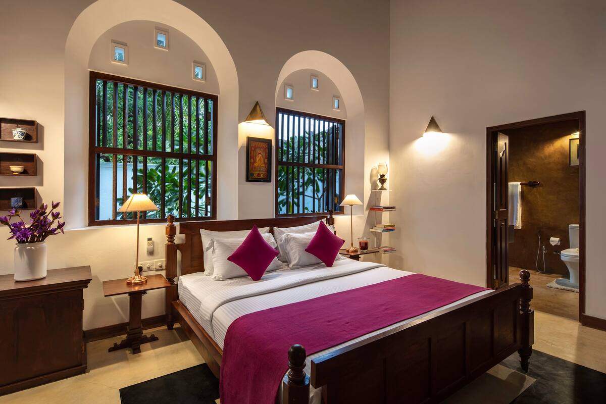 Un dormitorio con una cama grande, dos lámparas de noche, una mesita de noche con lámpara, una pequeña mesa con un jarrón de flores y una ventana con vistas a la vegetación.