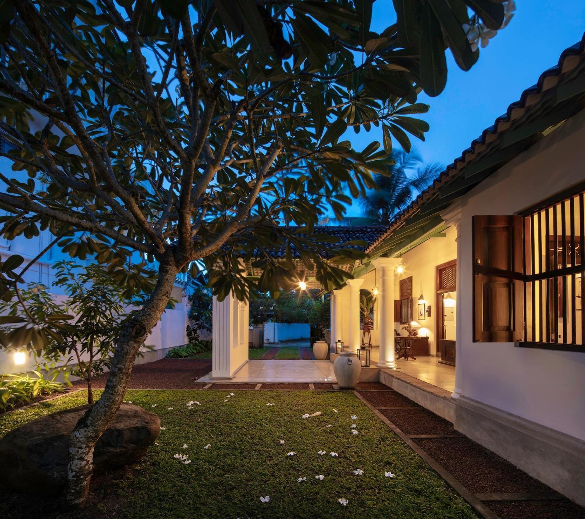 A well-lit garden with a pathway leading to a building with a tiled roof and white walls.