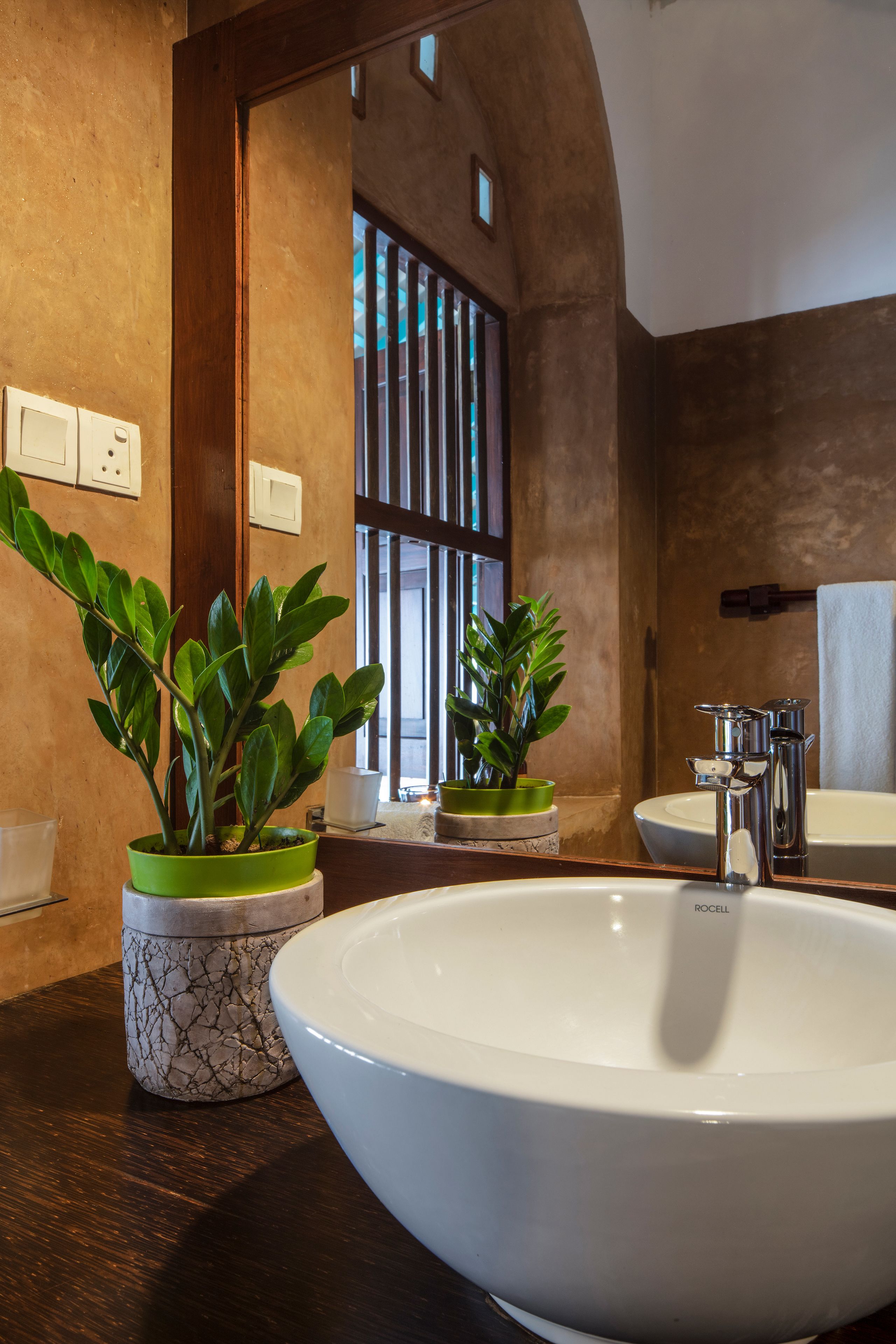 Un baño con un lavamanos grande de color blanco, dos plantas en macetas, un espejo y una ventana con persianas verticales.