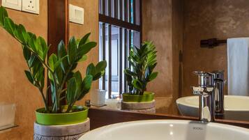 Un baño con un lavamanos grande de color blanco, dos plantas en macetas, un espejo y una ventana con persianas verticales.