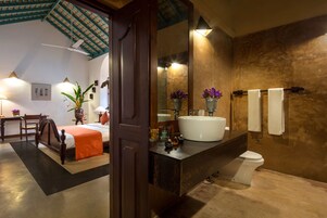 Un baño con un espejo grande, un lavabo y un inodoro. En la habitación contigua, hay una cama con una manta naranja y un cabecero de madera.