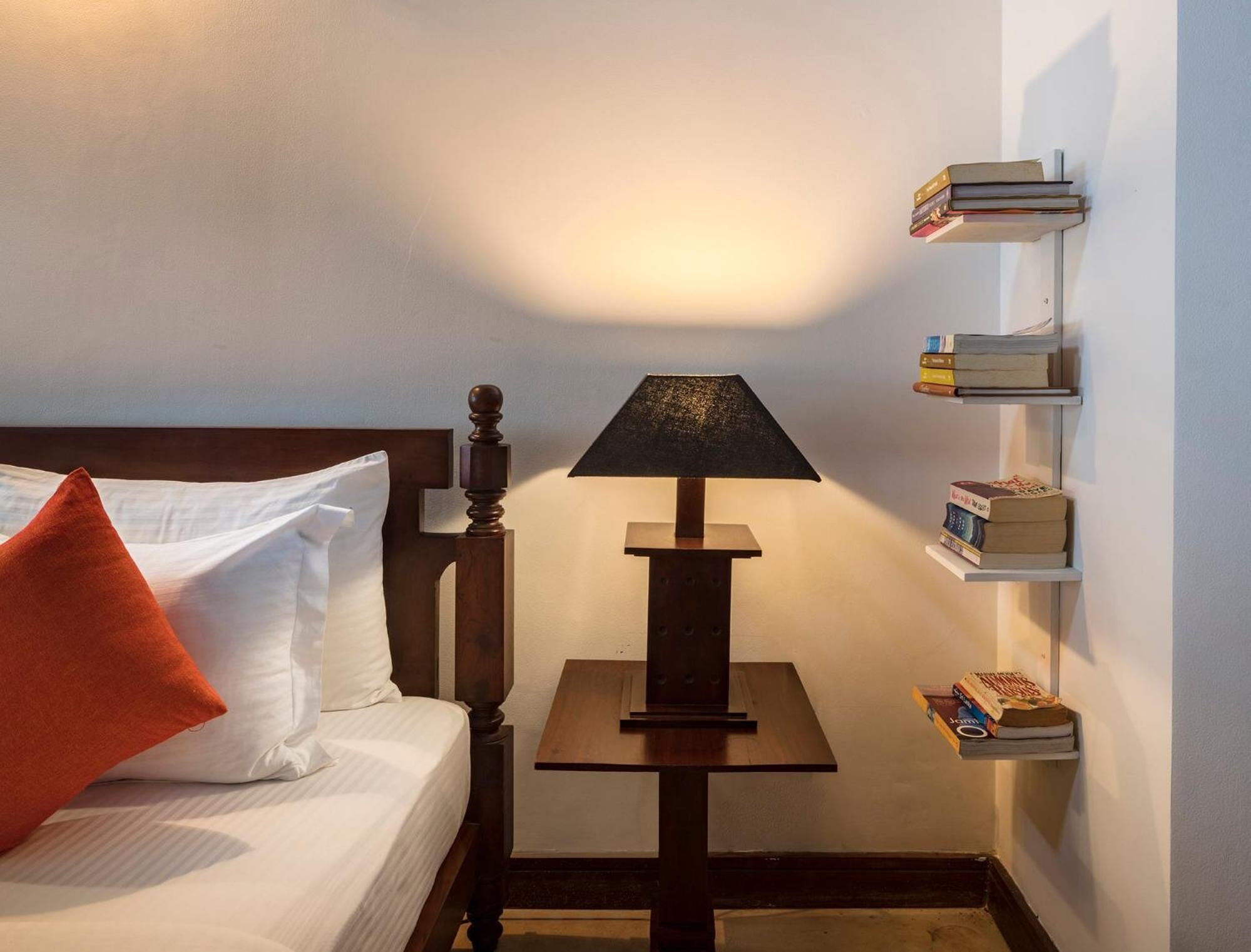 Una cama bien hecha con sábanas blancas y una almohada naranja, una mesita de madera con una lámpara, y una estantería con libros y un objeto decorativo.
