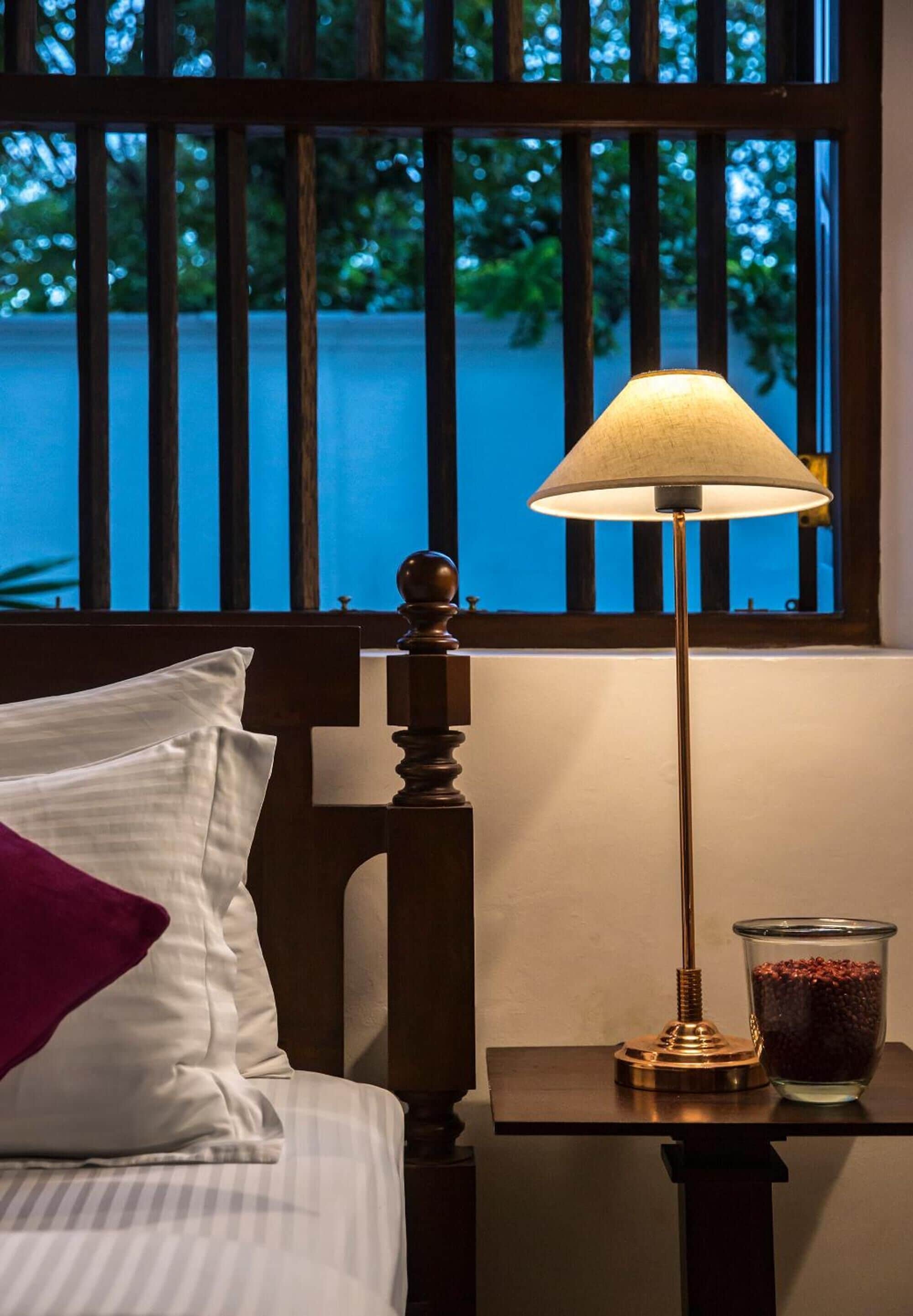 A bed with a white pillow and a purple accent pillow, a wooden headboard, a bedside table with a lamp and a glass container of snacks, and a window with vertical blinds overlooking a body of water.