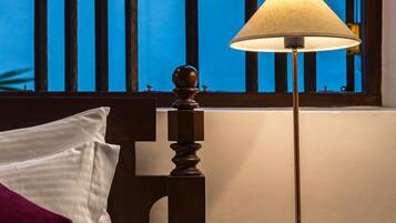 A bed with a white pillow and a purple accent pillow, a wooden headboard, a bedside table with a lamp and a glass container of snacks, and a window with vertical blinds overlooking a body of water.