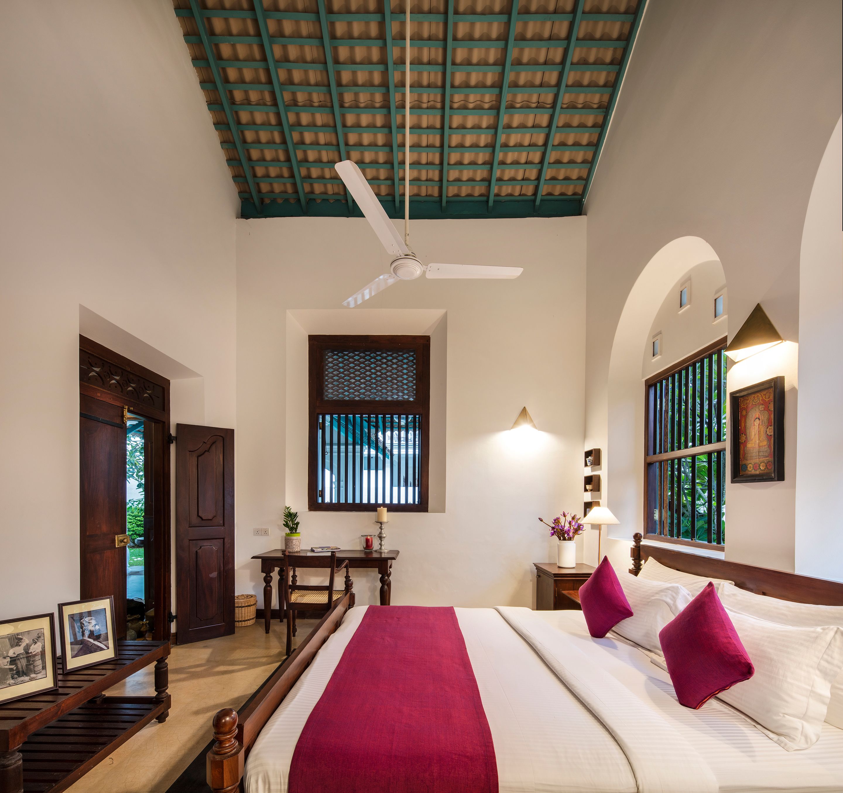 A hotel room with a bed, a desk, a chair, a ceiling fan, and a window with bars.