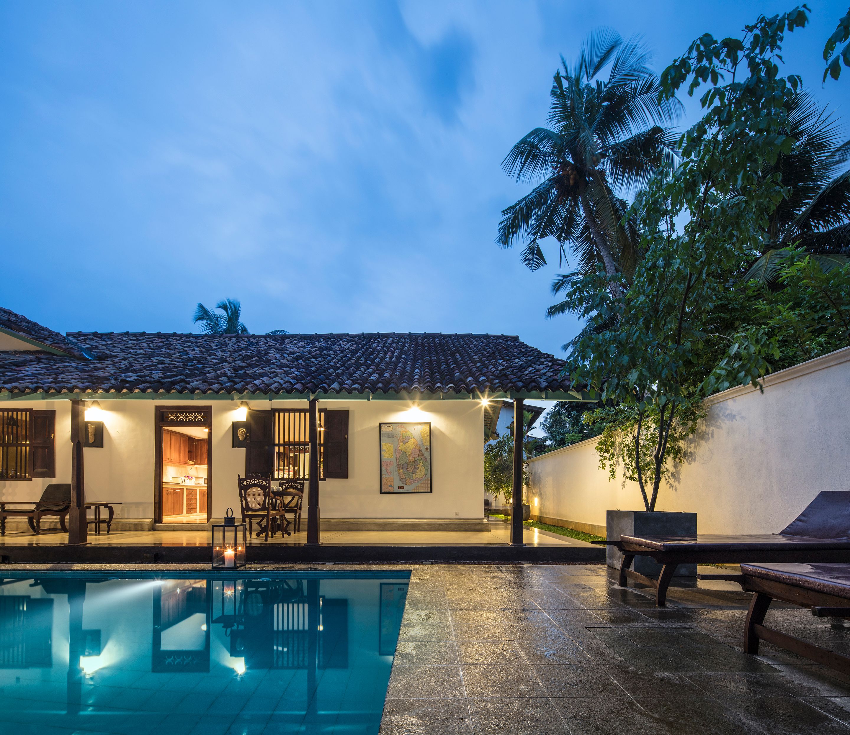 A poolside area with a tiled floor, a pool, and a building with a tiled roof and a door leading to a room.