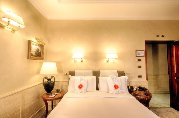 Quarto de hotel com uma cama grande, duas luminárias de cabeceira, uma mesa de cabeceira com abajur, um quadro na parede e uma porta de madeira.