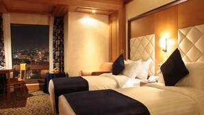 A hotel room with two beds, a desk with a chair, a small table with fruit, and a city view through the window. - Thousand Nights Hotel Amman (Amman)
