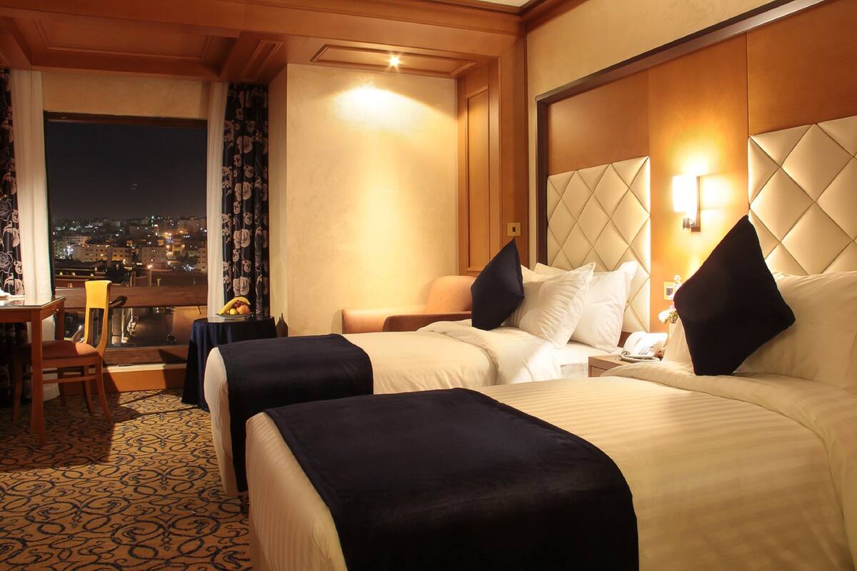 A hotel room with two beds, a desk with a chair, a small table with fruit, and a city view through the window.