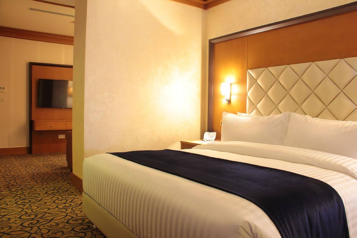 A hotel room with a large bed, a nightstand with a lamp, a television on a stand, and a patterned carpet.