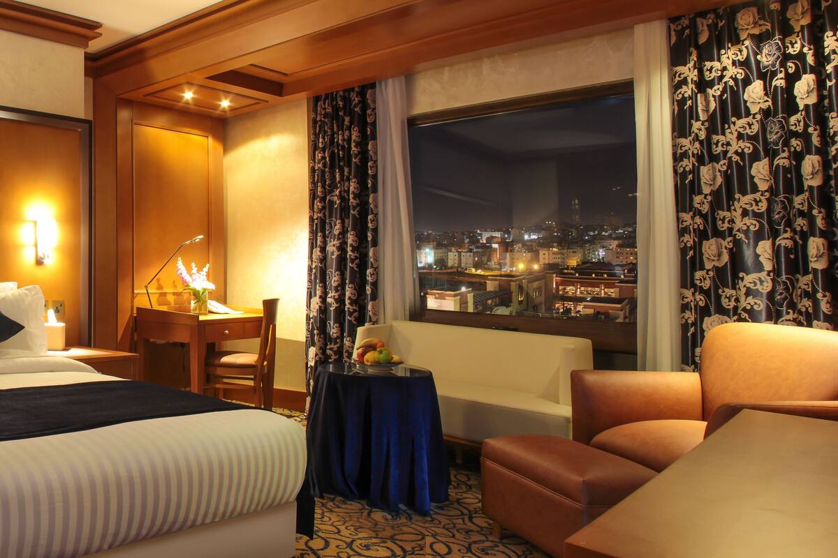 A hotel room with a large bed, a desk, a chair, and a view of the city at night.
