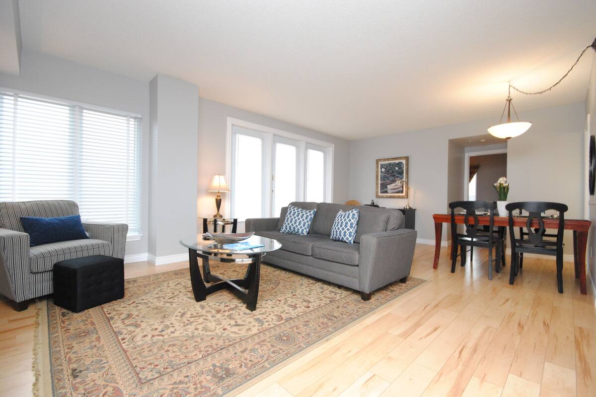 A living room with a gray sofa, a coffee table, a patterned rug, and a dining area with a wooden table and chairs.