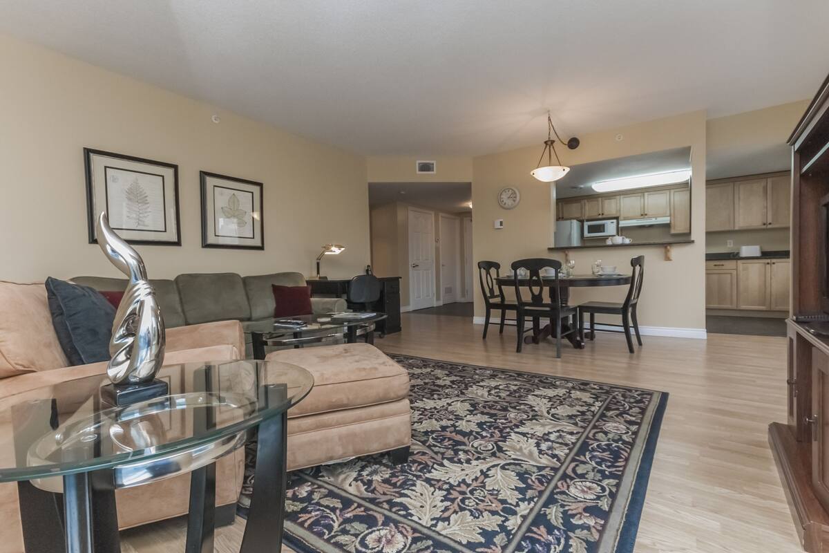 A living room with a glass coffee table, a beige sofa, and a patterned rug. There is a dining area with a table and chairs, and a kitchen in the background.