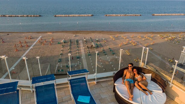 A balcony with lounge chairs, a wicker sofa, and a view of the beach with umbrellas and sun loungers. - Hotel International (Gabicce Mare)