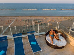 A balcony with lounge chairs, a wicker sofa, and a view of the beach with umbrellas and sun loungers. - Hotel International (Gabicce Mare)