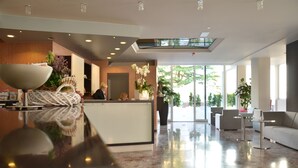 A modern hotel lobby with a reception desk, seating area, and large windows offering a view of the outdoors. - Hotel Amburgo (San Michele al Tagliamento)
