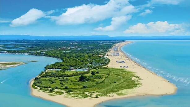 An aerial view of a coastal area with a beach, a small island with greenery, and a clear blue sky. - Hotel Amburgo (San Michele al Tagliamento)