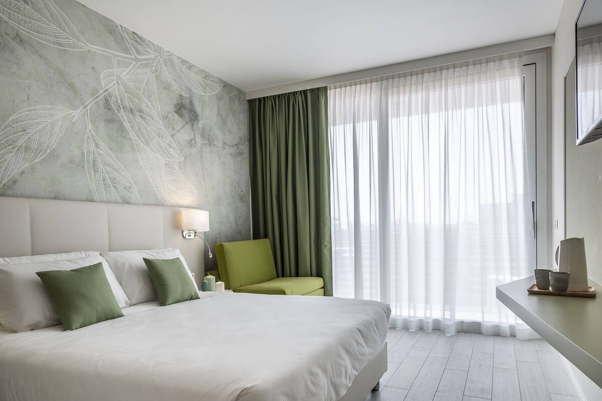 Et moderne hotelværelse med en stor seng, en grøn lænestol, et natbord med lampe og lette gardiner foran et vindue.