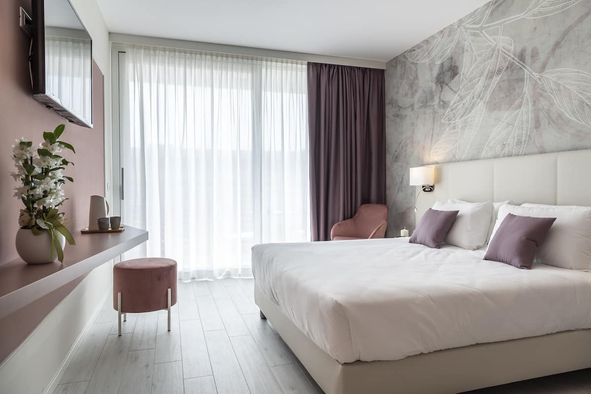 Et moderne hotelværelse med en stor seng, et sminkebord med spejl, en lille rund skammel, en stol og en dekorativ væg.