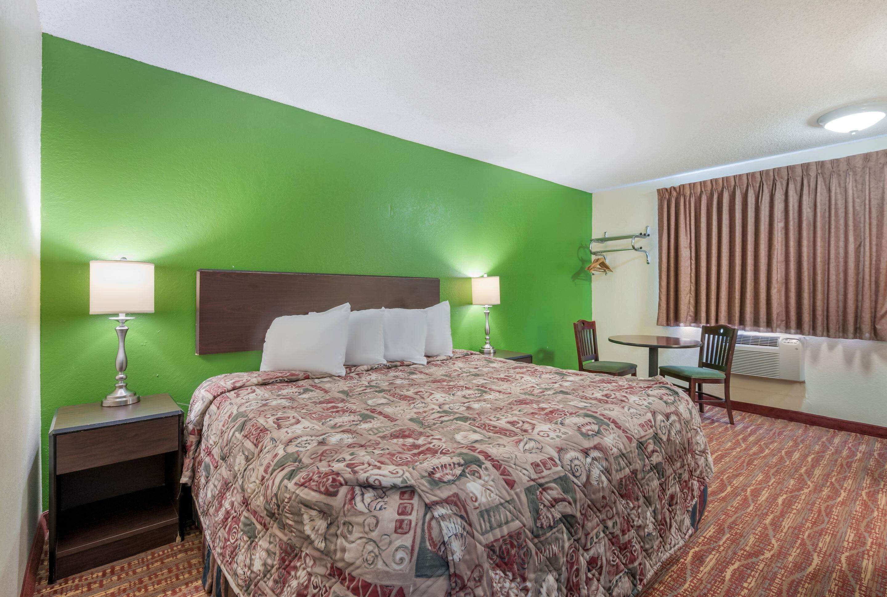 A hotel room with a green wall, a bed with a patterned bedspread, two bedside lamps, a small table with a chair, and a window with curtains.
