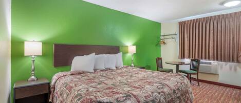 Une chambre d’hôtel avec un mur vert, un lit recouvert d’une couvre-lit à motifs, deux lampes de chevet, une petite table avec une chaise et une fenêtre avec des rideaux.