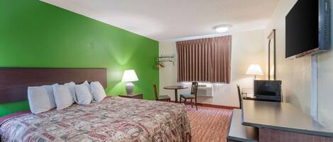 A hotel room with a large bed, a desk with a flat-screen TV, a chair, a table, and a window with curtains.