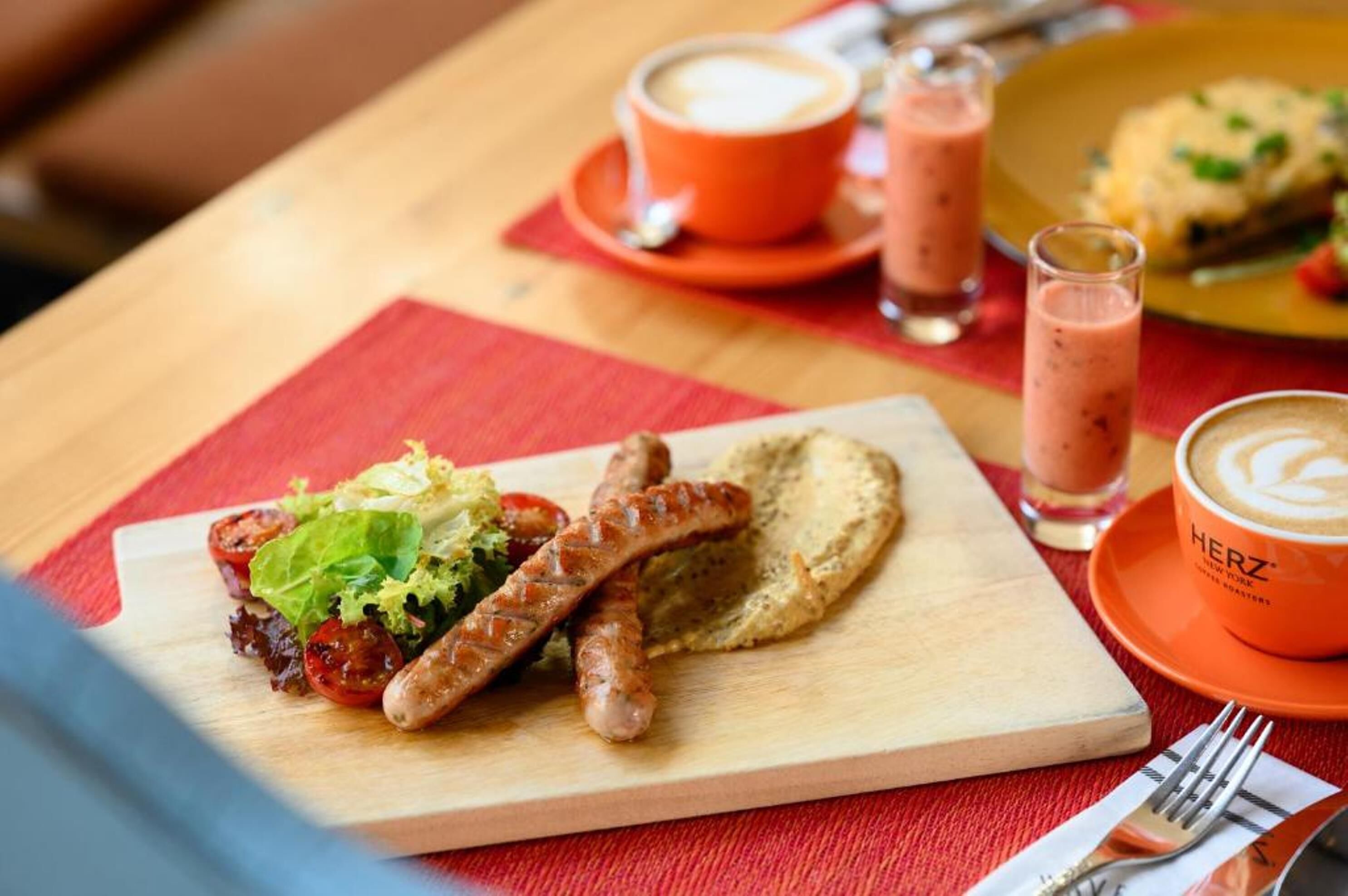 Un plato con salchichas, ensalada y una rebanada de pan sobre una tabla de madera, acompañado de una taza de café y un vaso de una bebida rosada sobre un mantel rojo.