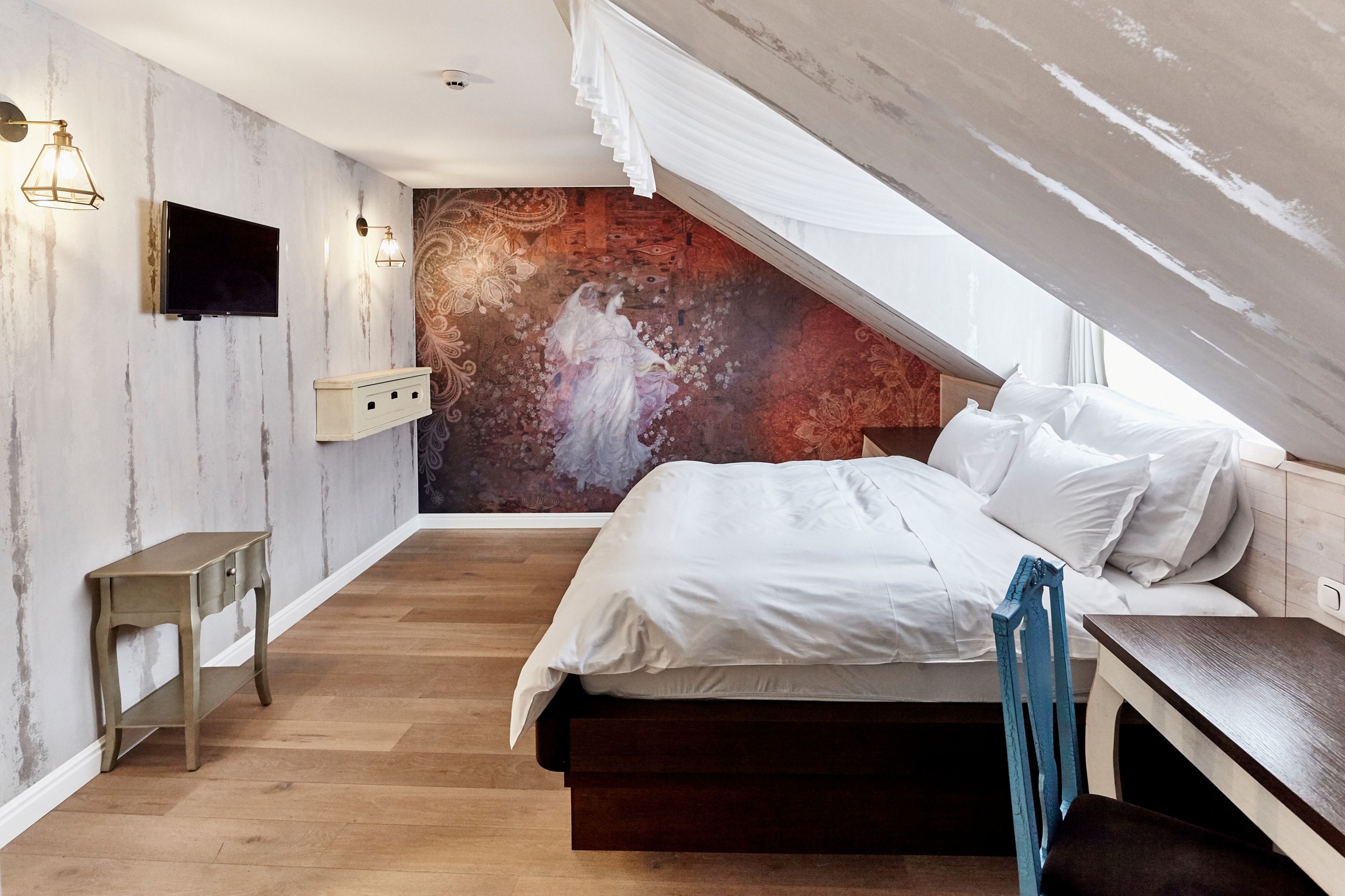 A bedroom with a wooden floor, a bed with white bedding, a TV mounted on the wall, and a decorative mural on the wall.