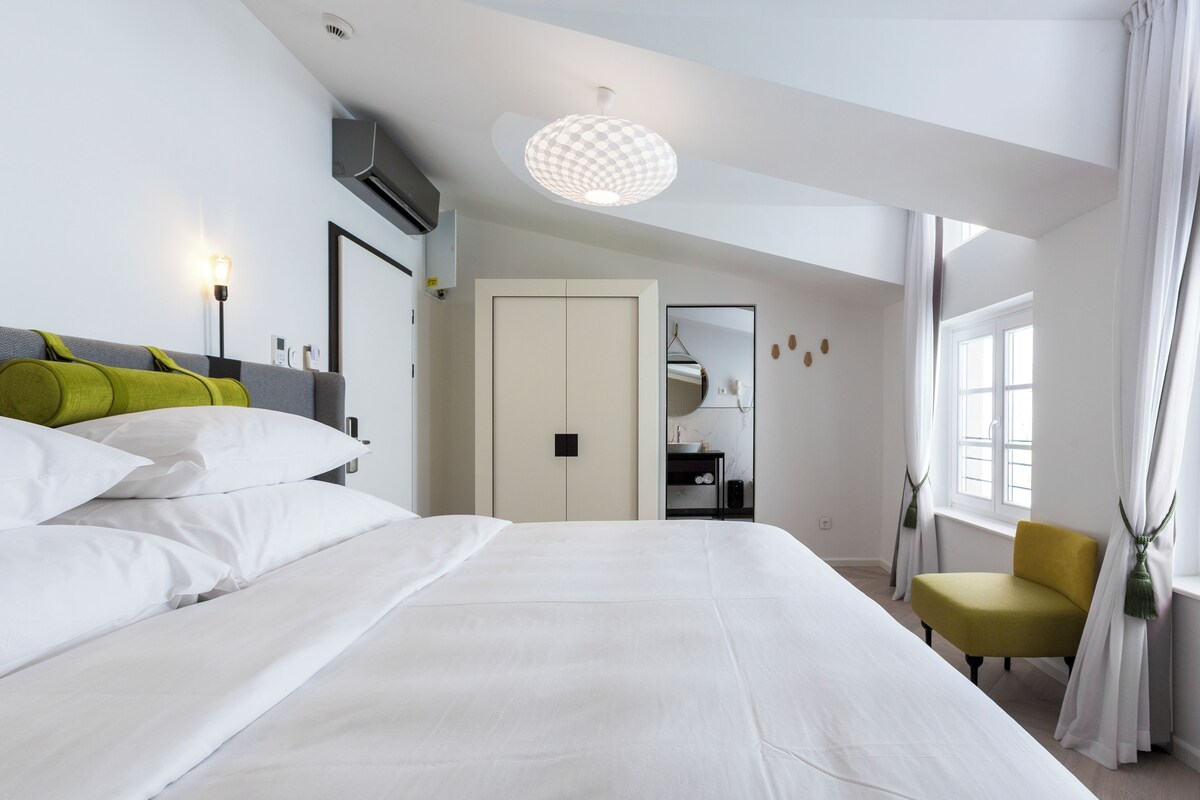 Une chambre d’hôtel moderne avec un grand lit, un fauteuil jaune, une armoire et une fenêtre avec des rideaux.