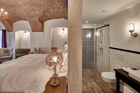A bedroom with a large bed, a decorative lamp, and a view of a bathroom with a shower and a toilet. - Maison Bistro & Hotel (Budapest)