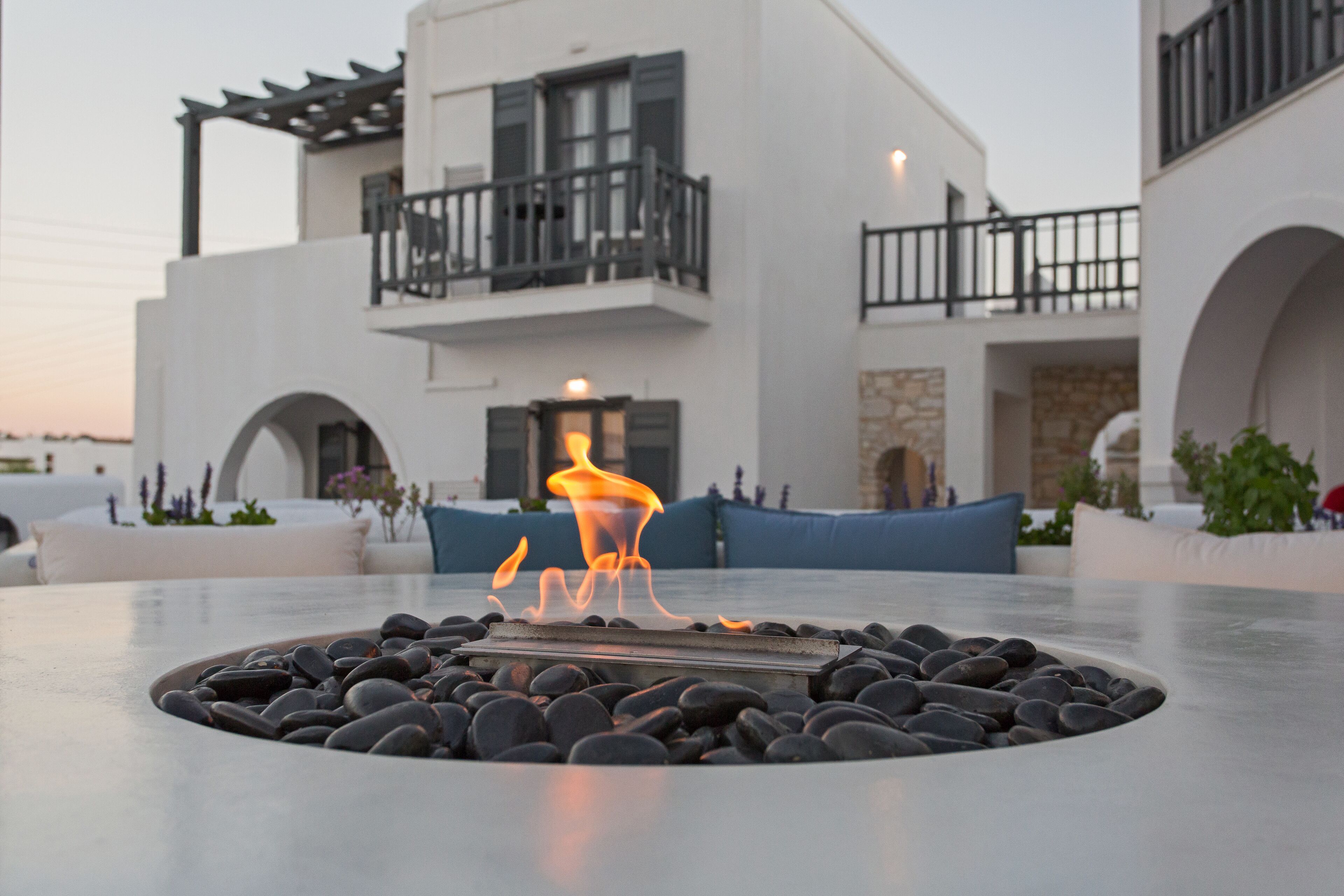 A modern outdoor seating area with a fire feature, surrounded by black stones, and a white building with balconies in the background.