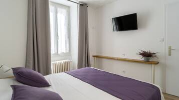Quarto com cama, cortinas, televisão na parede, radiador e estante com uma planta.