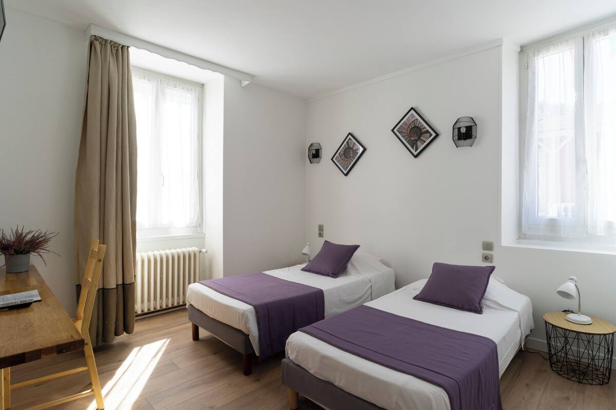 Una habitación con dos camas, una mesa de madera, una silla, un radiador y una ventana con cortinas.